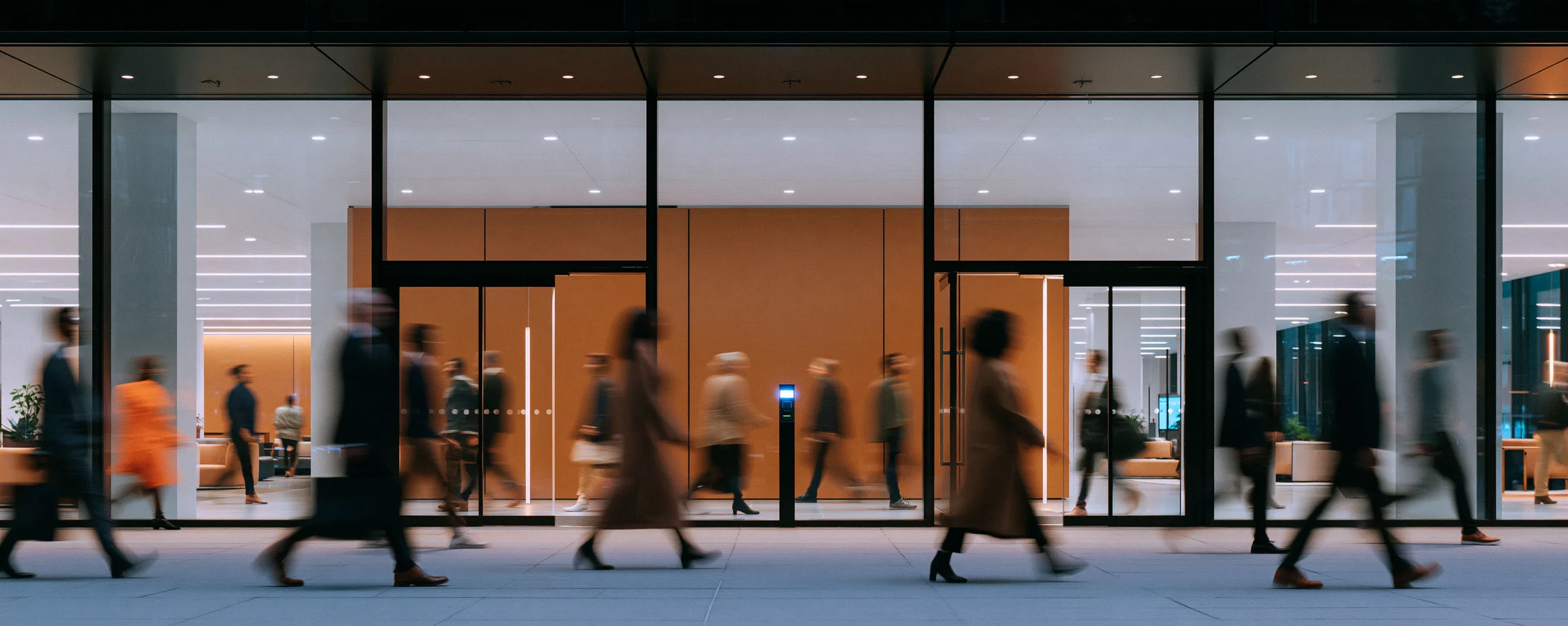 People walking past and through a glass office entrance at dusk, blurred in motion, a small lit badge reader by the doors that nobody stops at.